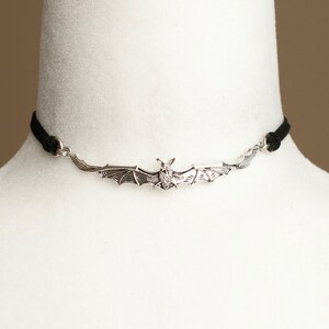 May include: A silver-toned metal bat pendant centered on a thin black cord choker. The bat is depicted with wings spread wide, showing detailed wing textures and a small head. The pendant is attached to the cord via small silver-toned metal rings.