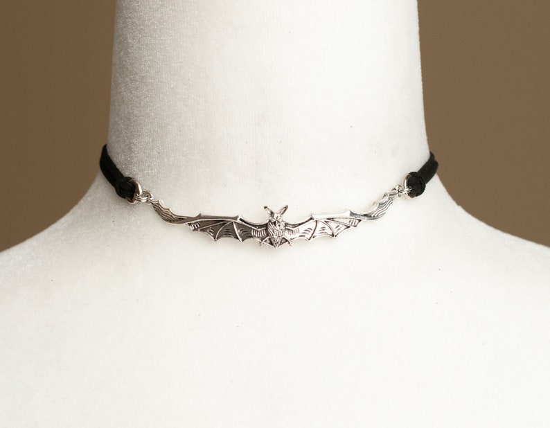 May include: A silver-toned metal bat pendant centered on a thin black cord choker. The bat is depicted with wings spread wide, showing detailed wing textures and a small head. The pendant is attached to the cord via small silver-toned metal rings.