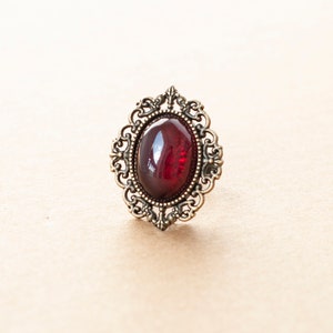 May include: A vintage-style ring with a dark red oval-shaped gemstone set in an ornate gold-toned metal frame.