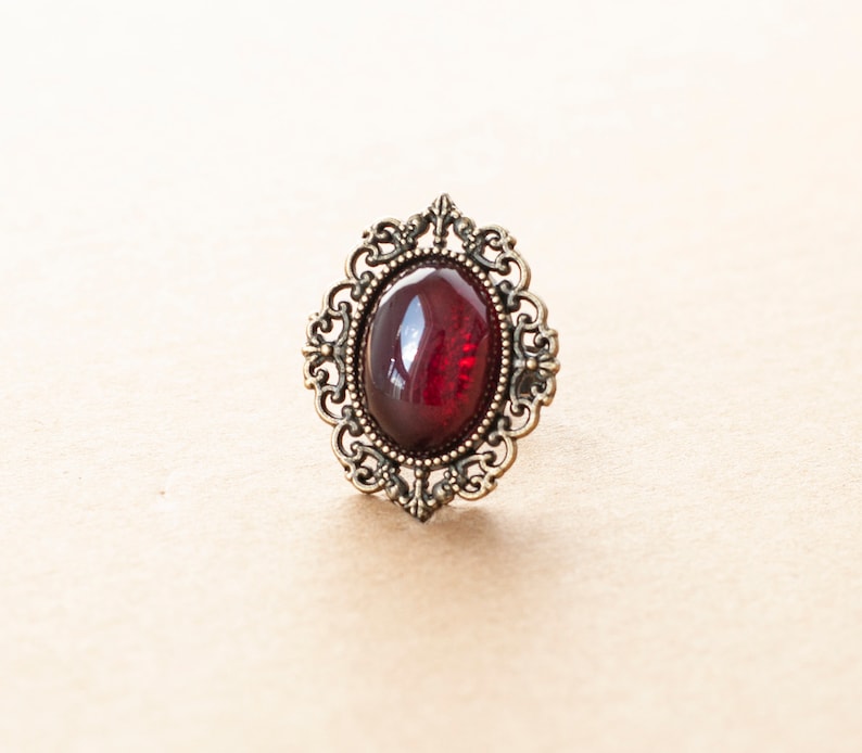 May include: A vintage-style ring with a dark red oval-shaped gemstone set in an ornate gold-toned metal frame.