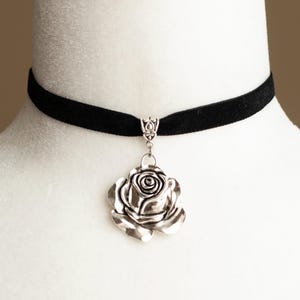 Puede incluir: Un collar tipo gargantilla de terciopelo negro con un colgante de rosa de metal plateado. El colgante está unido a la banda con un pasador decorativo de tono plateado. La rosa tiene pétalos detallados y superpuestos con un acabado pulido.