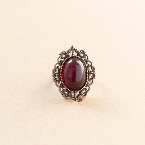 May include: A vintage-style ring with a dark red oval-shaped gemstone set in an ornate gold-toned metal frame.