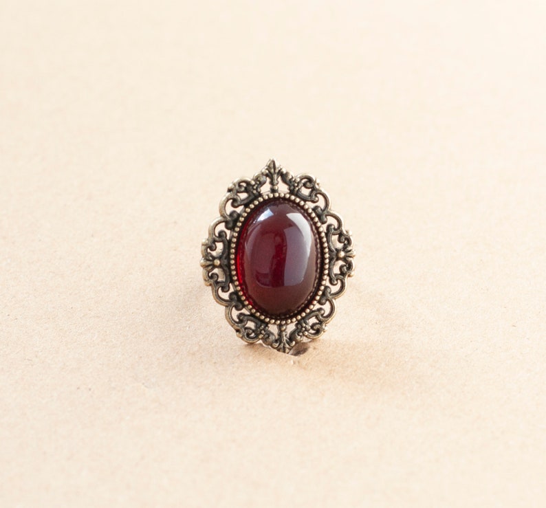 May include: A vintage-style ring with a dark red oval-shaped gemstone set in an ornate gold-toned metal frame.