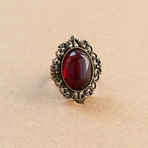 May include: A vintage-style ring with a large, oval-shaped, dark red gemstone set in an ornate, gold-toned metal frame.
