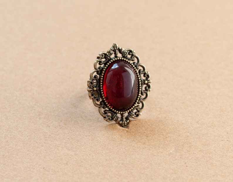May include: A vintage-style ring with a large, oval-shaped, dark red gemstone set in an ornate, gold-toned metal frame.