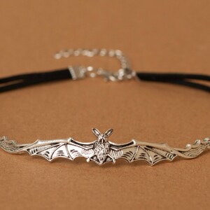 May include: A black velvet choker necklace featuring a central silver-toned metal bat pendant with outstretched wings. The necklace includes a silver-toned metal lobster claw clasp and an adjustable extender chain at the back for a secure fit.