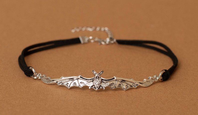 May include: A black velvet choker necklace featuring a central silver-toned metal bat pendant with outstretched wings. The necklace includes a silver-toned metal lobster claw clasp and an adjustable extender chain at the back for a secure fit.