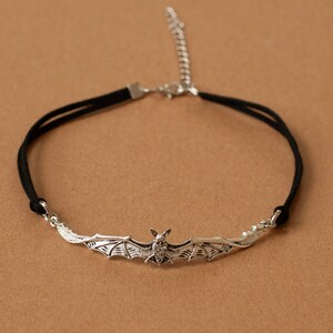 May include: A black cord choker necklace featuring a silver-toned metal bat pendant with outstretched wings. The necklace includes a silver-toned metal lobster claw clasp and an adjustable extender chain for a secure and customizable fit.