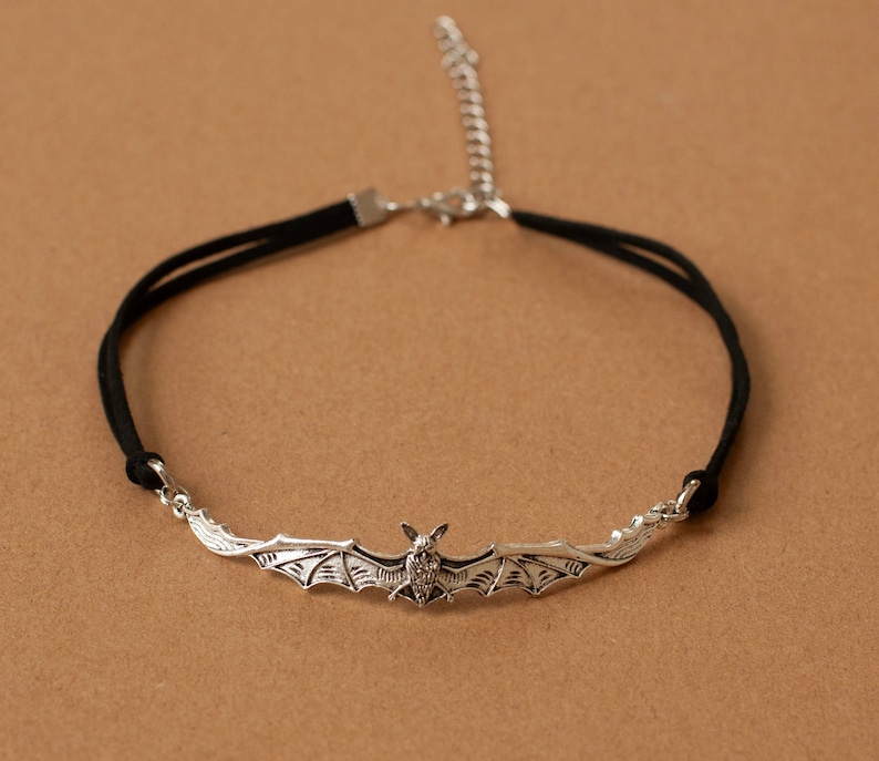 May include: A black cord choker necklace featuring a silver-toned metal bat pendant with outstretched wings. The necklace includes a silver-toned metal lobster claw clasp and an adjustable extender chain for a secure and customizable fit.