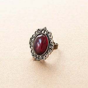 May include: A ring with a large, oval-shaped, dark red gemstone set in an ornate, gold-toned metal frame.