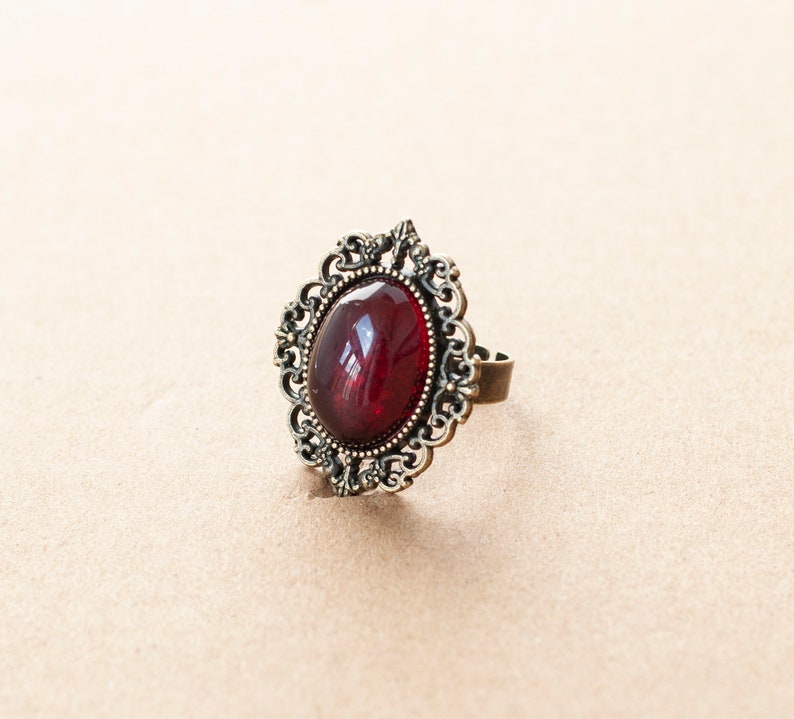 May include: A ring with a large, oval-shaped, dark red gemstone set in an ornate, gold-toned metal frame.