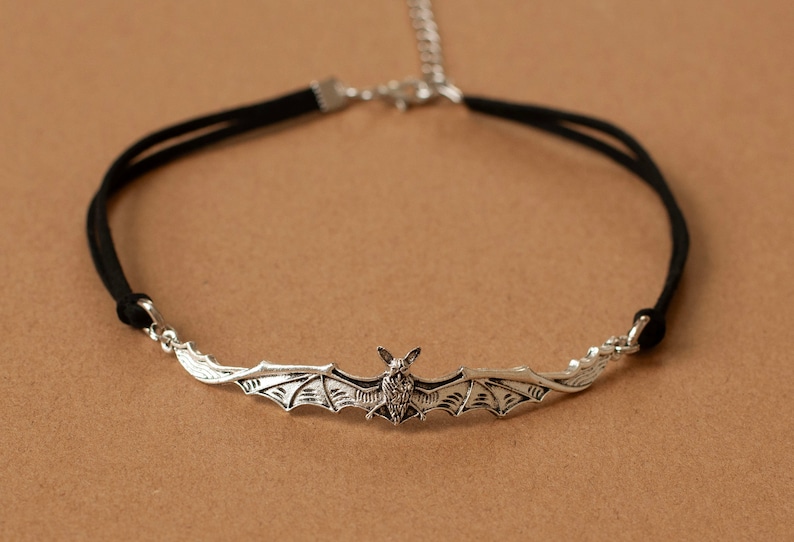 May include: A black cord choker necklace featuring a silver-toned metal bat pendant with outstretched wings. The necklace includes a silver-toned metal chain extender and a lobster claw clasp, providing an adjustable fit for the wearer.