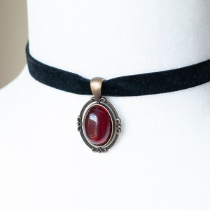 Puede incluir: Un collar tipo gargantilla de terciopelo negro con un colgante ovalado que presenta una piedra cabuj&oacute;n de color rojo intenso, engastada en un marco de metal color bronce antiguo con detalles decorativos de volutas en los bordes.