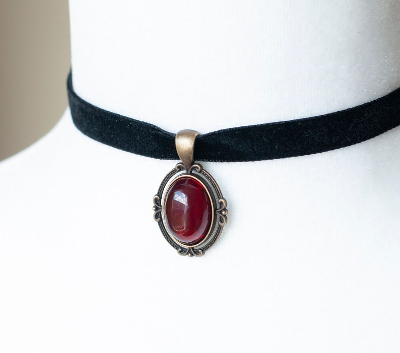 Puede incluir: Un collar tipo gargantilla de terciopelo negro con un colgante ovalado que presenta una piedra cabuj&oacute;n de color rojo intenso, engastada en un marco de metal color bronce antiguo con detalles decorativos de volutas en los bordes.