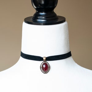 Puede incluir: Un collar tipo gargantilla de terciopelo negro con un colgante ovalado central. El colgante tiene una piedra cabuj&oacute;n de color rojo intenso engastada en un marco de metal de tono antiguo con detalles decorativos de volutas en los bordes.