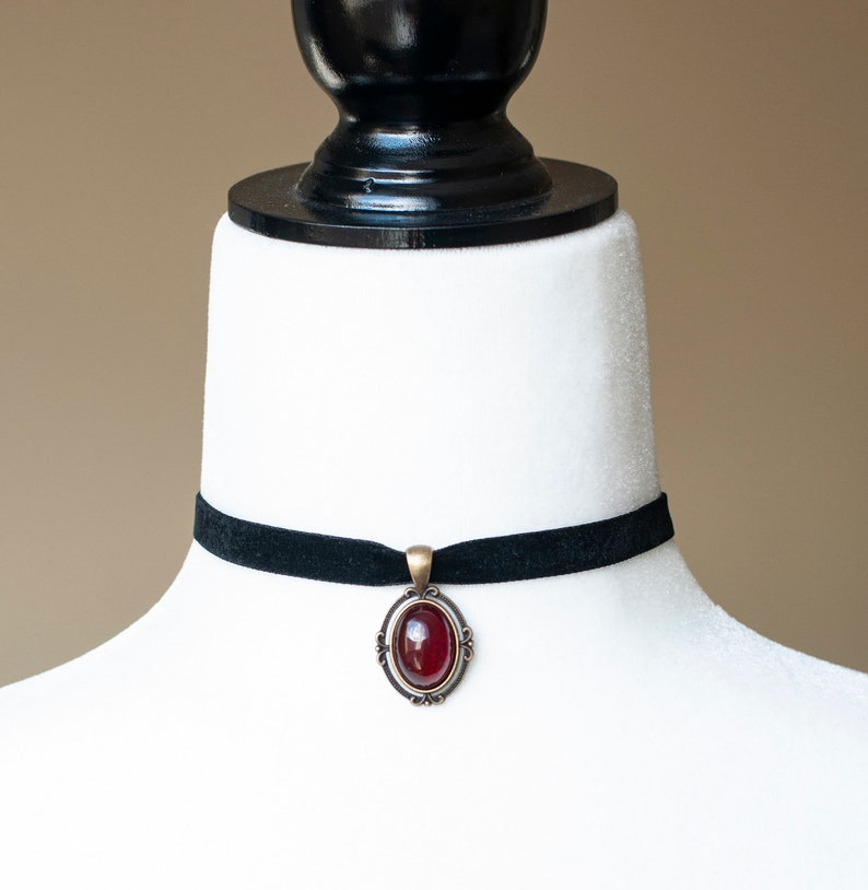 Puede incluir: Un collar tipo gargantilla de terciopelo negro con un colgante ovalado central. El colgante tiene una piedra cabuj&oacute;n de color rojo intenso engastada en un marco de metal de tono antiguo con detalles decorativos de volutas en los bordes.