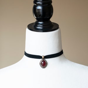 Puede incluir: Un collar tipo gargantilla de terciopelo negro con un colgante ovalado central que presenta una piedra cabuj&oacute;n de color rojo intenso, engastada en un marco de metal color bronce antiguo con detalles de filigrana en los bordes.