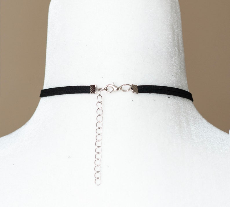 May include: A black choker necklace featuring a thin band, silver-toned metal crimp ends, a lobster claw clasp, and a dangling silver-toned metal extender chain with circular links, shown from the back view on a white display form.