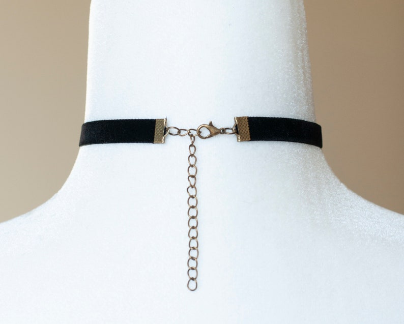 Puede incluir: Un collar tipo gargantilla de terciopelo negro con terminales de metal en tono bronce, un cierre de mosquet&oacute;n y una cadena extensora ajustable. El collar se muestra en el cuello de un maniqu&iacute; blanco, revelando el mecanismo de cierre.