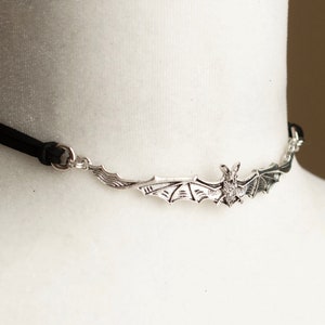 May include: A black choker necklace featuring a central silver-toned metal bat pendant with outstretched wings. The bat is attached to a thin black cord on both sides by small silver-toned metal rings, creating a horizontal design across the neck.
