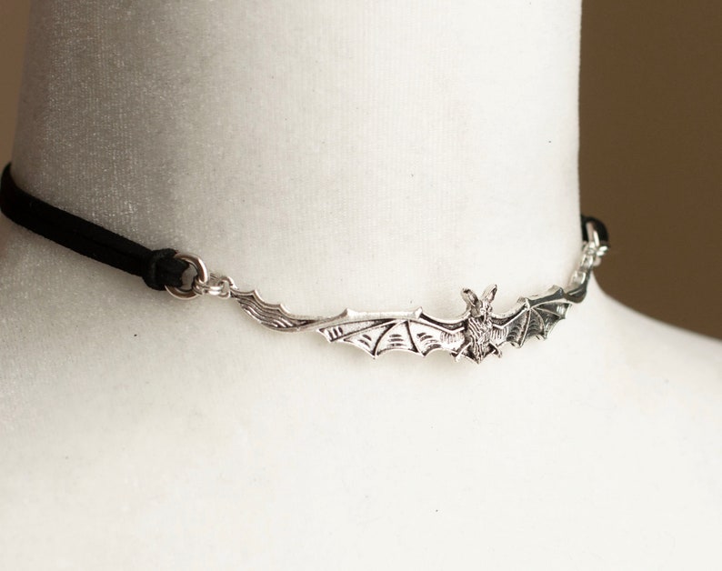 May include: A black choker necklace featuring a central silver-toned metal bat pendant with outstretched wings. The bat is attached to a thin black cord on both sides by small silver-toned metal rings, creating a horizontal design across the neck.