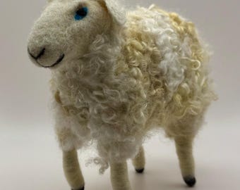 May include: A handcrafted sheep figurine with a fluffy, cream-colored wool body and a smiling face. The sheep has blue eyes and black hooves. The wool is a mix of white and light yellow.
