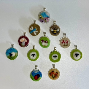 Könnte beinhalten: Eine Sammlung handgefertigter Filzanhänger mit silberfarbenen Ösen. Die Anhänger zeigen verschiedene von der Natur inspirierte Designs, darunter eine Libelle, eine Biene, Pilze, ein Schaf und ein farbenfrohes abstraktes Design. Der Filz ist in Grün-, Braun-, Blau- und Rottönen gehalten.