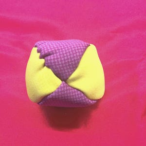 Puede incluir: Un pequeño juguete de tela hecho a mano con forma de cubo. El cubo está compuesto de tela morada y amarilla. La tela morada tiene un pequeño estampado de cuadros. La tela amarilla es de color liso. El juguete está probablemente diseñado para niños.