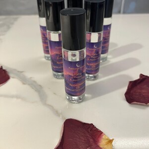 May include: Several small glass bottles with black lids are arranged on a white surface. The bottles have labels with a purple and orange sunset design and the text "Fantasy Island". Dried rose petals are scattered around the bottles.