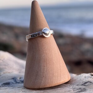 May include: A silver ring with a textured band and a rounded, raised design. The ring is displayed on a wooden cone-shaped stand. The background shows a blurred view of the sea and sky.