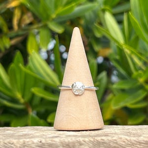 May include: A silver ring with a textured, round gemstone is displayed on a wooden cone. The ring's band is thin and delicate, and the gemstone has a faceted appearance. The background features green foliage, suggesting an outdoor setting.