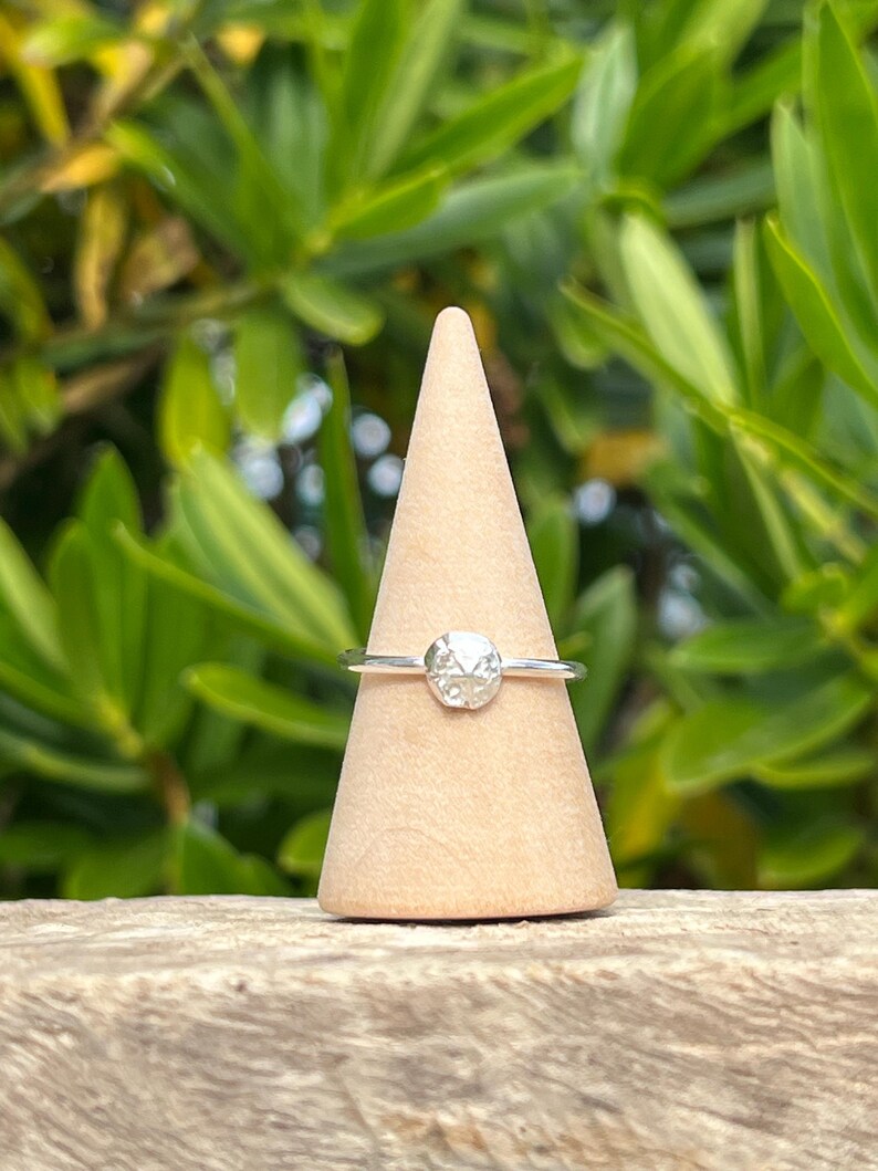 May include: A silver ring with a textured, round gemstone is displayed on a wooden cone. The ring's band is thin and delicate, and the gemstone has a faceted appearance. The background features green foliage, suggesting an outdoor setting.