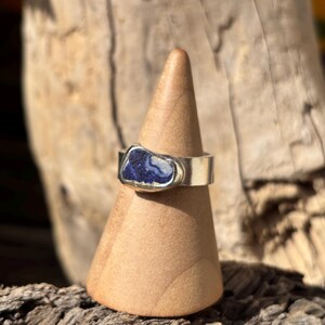 May include: A silver ring featuring a dark blue stone with white marbling. The ring is presented on a wooden, cone-shaped stand. The background shows blurred wood.