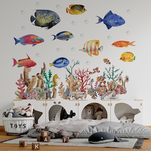 Può includere: Un set di adesivi murali con pesci colorati ed elementi di barriera corallina. Il design include varie specie di pesci nei toni del blu, giallo, arancione e rosso, insieme a coralli e bolle. Gli adesivi sono disposti sopra un mobile contenitore bianco con giocattoli.