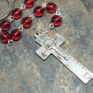 May include: A rosary keychain featuring a silver crucifix pendant with the inscription "INRI" and a figure of Jesus. The rosary beads are a deep red color and are connected by silver links. The keychain is designed for religious use.