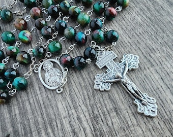May include: A rosary with green, black, and brown beads. The beads are strung on a silver chain, with a silver crucifix and a silver medallion featuring a religious figure. The rosary is displayed on a wooden surface.