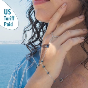 Könnte beinhalten: Eine silberne Handkette mit blauen Edelsteinakzenten. Die Kette verbindet einen Ring mit einem Armband, mit einem tropfenförmigen Edelstein in der Mitte der Hand. Weitere blaue Edelsteine sind entlang der Kette angeordnet. Der Text "US Tariff Paid" ist sichtbar.