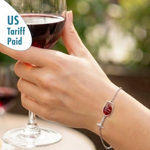 Könnte beinhalten: Ein silberfarbenes Kettenarmband mit einem zentralen Anhänger in Form eines Weinglases. Der Anhänger enthält eine tiefrote, durchscheinende Einlage, die Wein darstellt. Das Armband wird mit einer feinen Metallgliederkette am Handgelenk befestigt.
