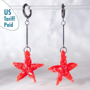 Könnte beinhalten: Ohrringe mit roten Seestern-Anhängern. Die Ohrringe haben silberfarbene Creolen und lange Stäbe. Die Seesterne sind strukturiert und an den Creolen aufgehängt. Ein auffälliges Accessoire.