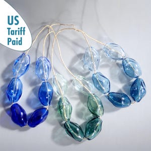 Könnte beinhalten: Drei Halsketten mit großen, oval geformten Glasperlen in Blau- und Grüntönen. Die Perlen sind auf einer dünnen, naturfarbenen Kordel aufgefädelt. Der Text "US Tariff Paid" befindet sich in der oberen linken Ecke.