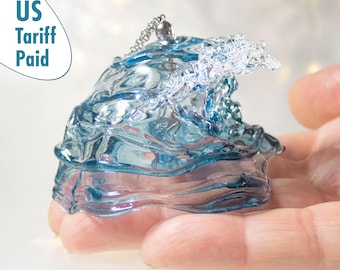 Peut inclure: Ornement en forme de vague bleue transparente avec une chaîne argentée. L'ornement est tenu dans une main. Le texte "US Tariff Paid" est dans le coin supérieur gauche. La vague est détaillée avec une surface texturée.