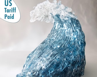 Peut inclure: Une sculpture décorative en verre représentant une vague, avec une crête en verre transparent et un corps en verre bleu. La vague est en mouvement, avec l'eau semblant se courber et se briser. Le texte "US Tariff Paid" est en haut à gauche.