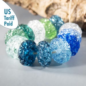 Puede incluir: Un collar de cuentas de vidrio de colores en tonos azules, verdes y transparentes. Las cuentas tienen un aspecto texturizado y agrietado. El texto "US Tariff Paid" es visible. Las cuentas están ensartadas.