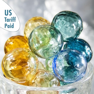 Könnte beinhalten: Eine Sammlung farbiger Glasmurmeln in Gelb-, Grün- und Blautönen. Die Murmeln werden in einem durchsichtigen Glasbehälter präsentiert. Das Bild enthält den Text "US Tariff Paid".