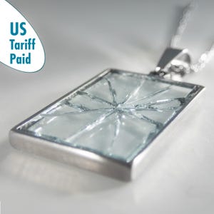 May include: A rectangular pendant necklace with a silver-colored frame. The pendant features a light blue, cracked glass design. The necklace is on a silver chain. The text "US Tariff Paid" is in the upper left corner.