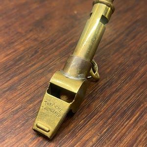 May include: A vintage brass whistle with the text "THE ACME THUNDERER" engraved on the front. The whistle has a cylindrical body and a small ring for attachment. The brass has a slightly aged appearance.