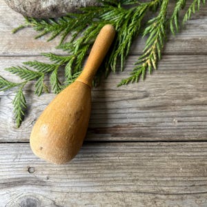 May include: A wooden mortar and pestle, with a light brown color and a smooth, worn surface. The pestle is lying on a wooden surface, with a green leafy branch in the background.