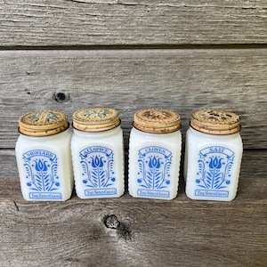 May include: Four vintage spice jars with white glass bodies and metal lids. Each jar has a blue floral design and the name of a spice: Mustard, Allspice, Cloves, and Salt. The text "The Spice Chest" is printed below the floral design.