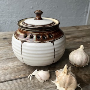May include: A white ceramic garlic keeper with a brown rim and lid, featuring small ventilation holes. The keeper is round with a lid and a small knob. Two whole garlic bulbs and a few cloves are next to the keeper on a wooden surface.