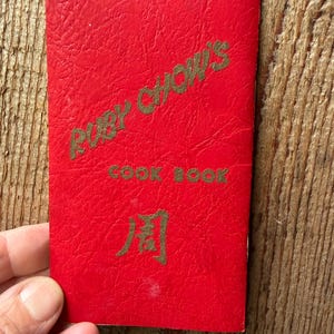 May include: A red cookbook titled "Ruby Chow's Cook Book" with gold lettering and a Chinese character. The cover has a textured appearance.
